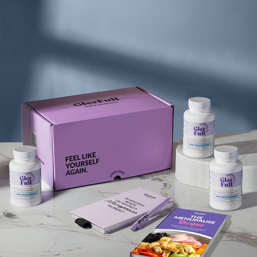 A product bundle display for GleeFull Supplements on a marble surface against a blue background. The layout includes a large purple shipping box featuring the slogan 'FEEL LIKE YOURSELF AGAIN.', three white bottles of Meno Belly Flusher, a purple branded pen, a matching notepad that reads 'REMINDERS WHEN I'M HAVING A MENOPAUSE MOMENT', and a recipe booklet titled 'THE MENOPAUSE Recipes'