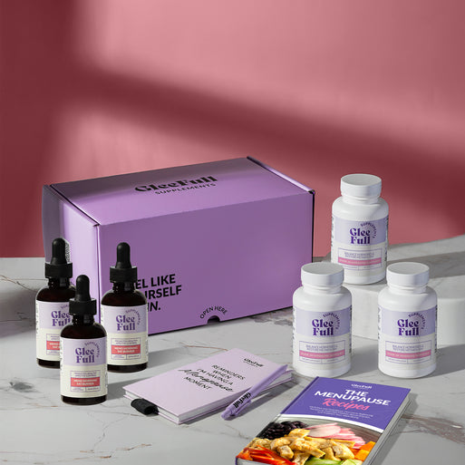 A product bundle display for GleeFull Supplements on a marble surface against a red background. The layout includes a large purple shipping box featuring the slogan 'FEEL LIKE YOURSELF AGAIN.', three white bottles of Over 30 Hormone Support and three black bottles of Meno Morning Fat Burner, a purple branded pen, a matching notepad that reads 'REMINDERS WHEN I'M HAVING A MENOPAUSE MOMENT', and a recipe booklet titled 'THE MENOPAUSE Recipes'