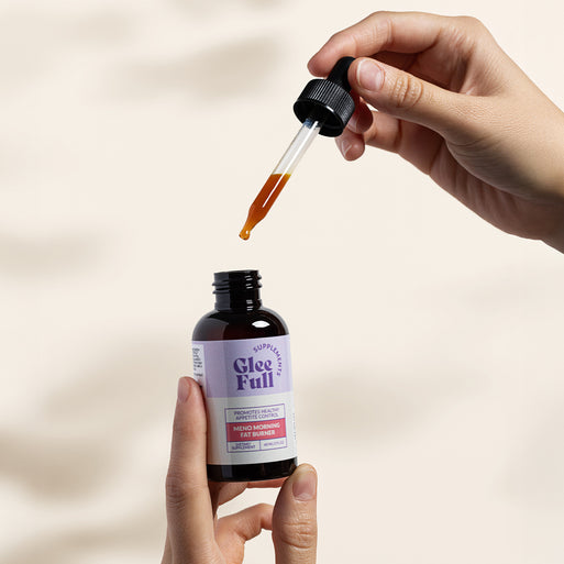 A person holds an amber GleeFull Meno Morning Fat Burner dropper bottle in one hand and the dropper in the other, releasing a drop of amber liquid, set against a soft cream background. The label reads 'Promotes Healthy Appetite Control, Meno Morning Fat Burner, 60 ML/2 FL OZ'.