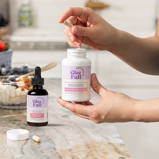 GleeFull Meno Morning Fat Burner (amber dropper) and Over 30 Hormone Support (white) bottles on a marble countertop in a kitchen setting.