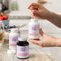 A person holds an Estro-Harmony bottle and a tan capsule in a kitchen, with Over 30 Hormone Support and Over 30 Hormone Support Plus bottles on a marble countertop alongside fresh strawberries and a yogurt bowl.