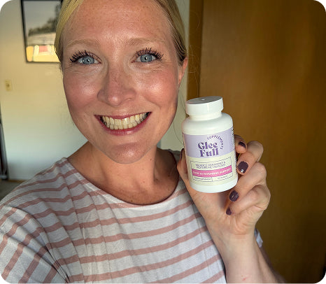 A smiling blonde woman holds up a white bottle of GleeFull dietary supplement in front of her face, wearing a pink and white striped shirt in an indoor setting.